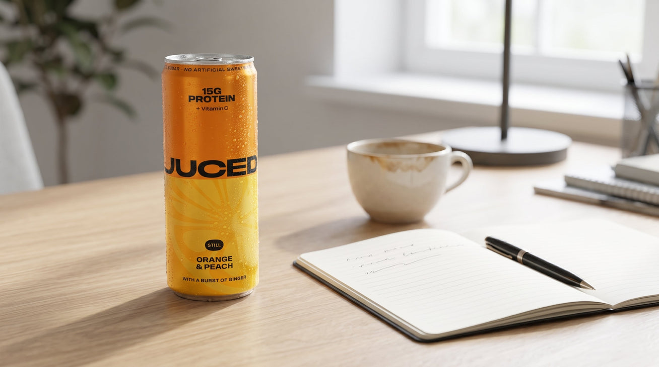 A can of JUCED fruit protein drink on a desk beside a notebook and morning coffee, illustrating protein's role in steady energy through the day