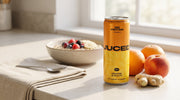 Can of JUCED Orange, Peach and Ginger protein drink on a kitchen counter next to a bowl of porridge, fresh oranges and ginger root, morning breakfast scene