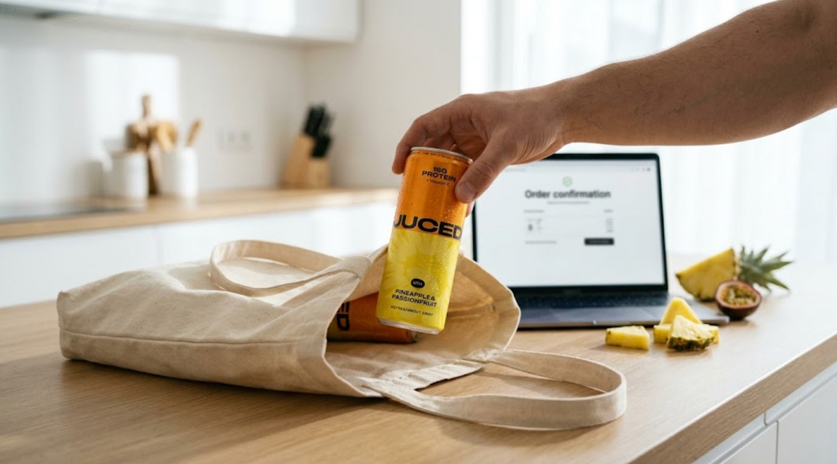 Hand placing a can of JUCED clean protein drink into a tote bag next to a laptop showing an online order, representing where to buy protein drinks in the UK