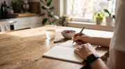 Hands writing a list in a notebook at a kitchen table